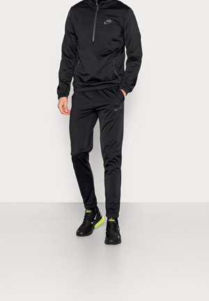 Mann trägt schwarzen Nike-Trainingsanzug mit Reißverschlussjacke und passenden Hosen, kombiniert mit schwarzen Nike-Sneakers mit neongelben Sohlen.