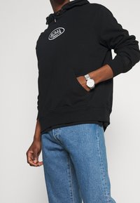 Svart huva sweatshirt med en framficka i kengurustil, prydd med ett vitt logopatch. Kombinerad med ljusblå jeans och en silverfärgad klocka.