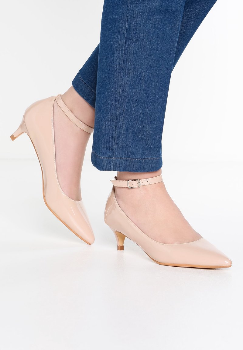Pieds chaussés de chaussures nude en cuir verni à talons petits avec brides autour de la cheville, associés à un pantalon en denim bleu sur fond blanc.