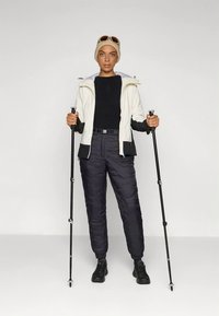 Viegls, izolēts balts jaciņa ar melnām uzsvariem, kombinēts ar melnām pūkaina stila biksēm. Tur pārgājienu nūjas, valkā bēša cepure un saulesbrilles.