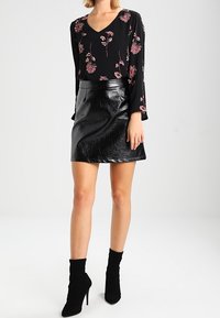 Blouse noire à imprimé floral avec manches longues et col en V, assortie à une mini jupe noire brillante, complétée par des bottines noires à talons.