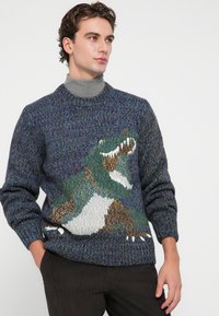 Pull en maille dans des tons de bleu et de vert, arborant un grand motif de dinosaure multicolore, avec un col rond et des poignets côtelés.