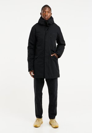 Sort isoleret parka med lynlås foran, justerbar hætte og forlommer. Båret over mørke jeans og gule sneakers.