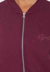 Gros plan sur une personne portant une veste zippée bordeaux avec un col côtelé et le mot partiel "Smile" imprimé sur le côté droit.