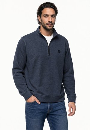 Mann mit dunklen Haaren und Bart, der einen marineblauen Pullover mit Viertelreißverschluss und blaue Jeans trägt, steht mit einer Hand in der Tasche vor einem einfarbigen Hintergrund.