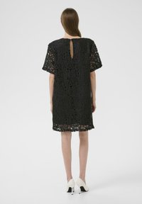 Rochie din dantelă neagră cu o croială lejeră, mâneci scurte și un decolteu în formă de lacăt la spate, având un tiv crestat și modele florale intricate.