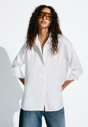 Göndör hajú nő nagy borostyánszínű napszemüvegben, laza, fehér, begombolható, felhajtott ujjú ingben és kék farmernadrágban, aki egy falnak támaszkodik.