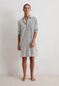 Mujer de pie descalza contra una pared blanca, vestida con un vestido de camisa de estampado blanco y negro y las mangas arremangadas.