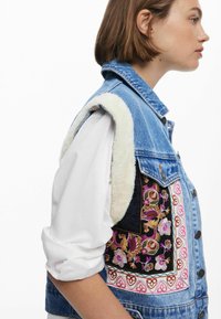 Denimweste mit Lammfellkragen, floralen Stickereien in Pink und Gold, mit Knopfverschluss und Kontraststoffdetails an den Seiten.