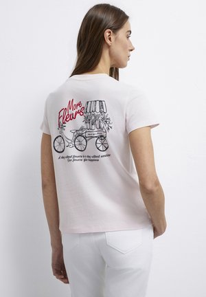 Woman in white pants and light pink T-shirt with a bicycle and flower cart graphic and red text "More Fleurs" on the back.