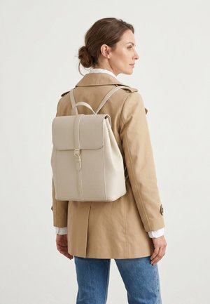 Vrouw met een beige trenchcoat en blauwe jeans draagt een lichtbeige leren rugzak met gespsluiting op haar rug.