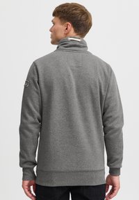 Pilka, tekstūruota megztinio tipo palaidinė su aukštu apykakle, raištuotomis rankogaliais ir apačia. Viršutinėje rankos dalyje yra siuvinėta logotipo detalė ir dekoratyvinė virvutė prie kaklo.