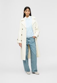 Femme portant un trench-coat blanc, un sweat-shirt bleu clair, une chemise blanche à col, un jean bleu ample et des chaussures blanches, debout devant un fond uni.