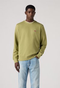 Sudadera verde oliva con cuello redondo y puños de canalé, que presenta un pequeño logo rojo en el pecho. Combinada con unos vaqueros de un azul claro.