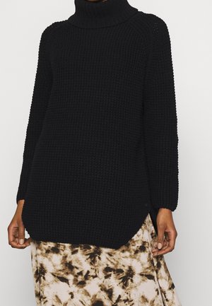 Person iført en sort tekstureret rullekrave sweater og et beige skørt med brune og sorte abstrakte mønstre.
