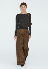 Persoon die een zwart en grijs gestreept langemouwen shirt draagt, brede bruine broek, zwarte schoenen en een bruine schoudertas draagt, staande tegen een witte muur.