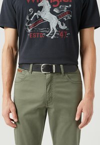 Svart t-shirt för män med grafiskt tryck och olivgröna byxor. Har ett svart bälte med en silverspänne. Slät tygstruktur.