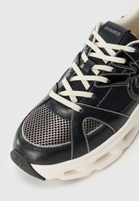 Baskets noires en mesh et cuir, avec des lacets blancs, une semelle crémeuse épaisse et des détails de logo sur la languette et le talon.