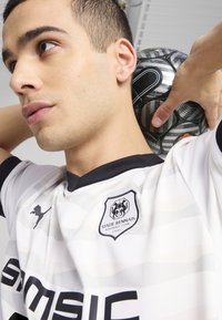 Biely futbalový dres s jemnými horizontálnymi vzormi, čiernymi akcentmi a logom Stade Rennais. Hráč drží štruktúrovanú čierno-sivú futbalovú loptu.