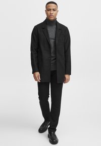 Abrigo largo negro sobre un suéter de cuello alto gris, combinado con pantalones negros ajustados y zapatos negros. Tela suave, diseño elegante y acentos sutiles.