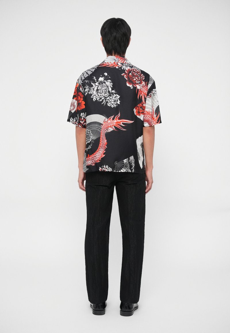 Kurzarmhemd aus schwarzem Stoff mit roten und weißen Blumen- und Drachenmustern, kombiniert mit schwarzen strukturierten Hosen und Schuhen.