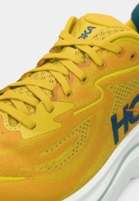 Κίτρινο αθλητικό παπούτσι με επάνω μέρος από δίχτυ, υφή με μοτίβο και μπλε λογότυπο. Διαθέτει επίπεδα κορδόνια και λευκή ενισχυμένη σόλα για υποστήριξη.