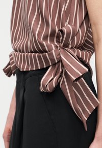 Καφέ και λευκή ριγέ μπλούζα δεμένη στο πλάι, συνδυασμένη με μαύρο παντελόνι ψηλής μέσης, σε κορμό και χέρι ατόμου.