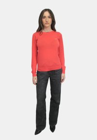 Maglione rosso a maniche lunghe con scollatura rotonda e polsini a costine, abbinato a jeans svasati in denim scuro, indossato con décolleté nere a punta.