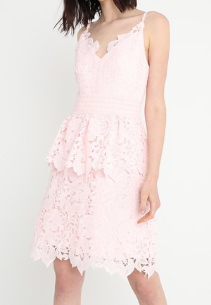 Vrouw die een lichtroze mouwloze kanten jurk draagt met bloemmotieven en een peplum taille detail tegen een effen achtergrond.