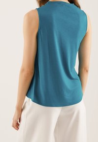 Haut sans manches couleur sarcelle en tissu doux avec un ourlet arrondi. Le dos présente un design simple, offrant une coupe décontractée. Assorti à un pantalon blanc.