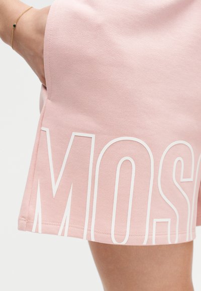 Κοντινό πλάνο ενός ατόμου που φοράει ροζ φούστα με μεγάλα λευκά γράμματα "MOS" τυπωμένα κοντά στο κάτω μέρος, με το χέρι να ακουμπά στην τσέπη και ένα χρυσό βραχιόλι στον καρπό.