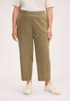Persoon draagt olijfgroene rechte broek met elastische tailleband, witte sneakers en een wit T-shirt met korte mouwen tegen een effen achtergrond.