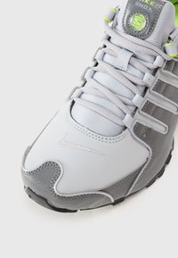 Sivi in beli Nike Shox copati z teksturirano usnjem, kovinskimi akcenti in mrežastim jezikom. Imajo odsevniki logo in svetlo zelene podrobnosti.