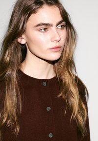 Brązowy sweter z dzianiny o okrągłym dekolcie i ciemnych guzikach. Modelka ma długie, falujące włosy, minimalny makijaż i neutralne tło.