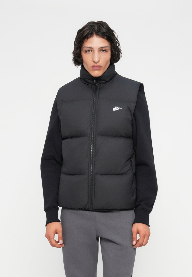 Jovem com cabelo encaracolado e escuro, vestindo um colete acolchoado preto da Nike sobre uma camisa de manga longa preta e calças de moletom cinzas, de pé contra um fundo branco.