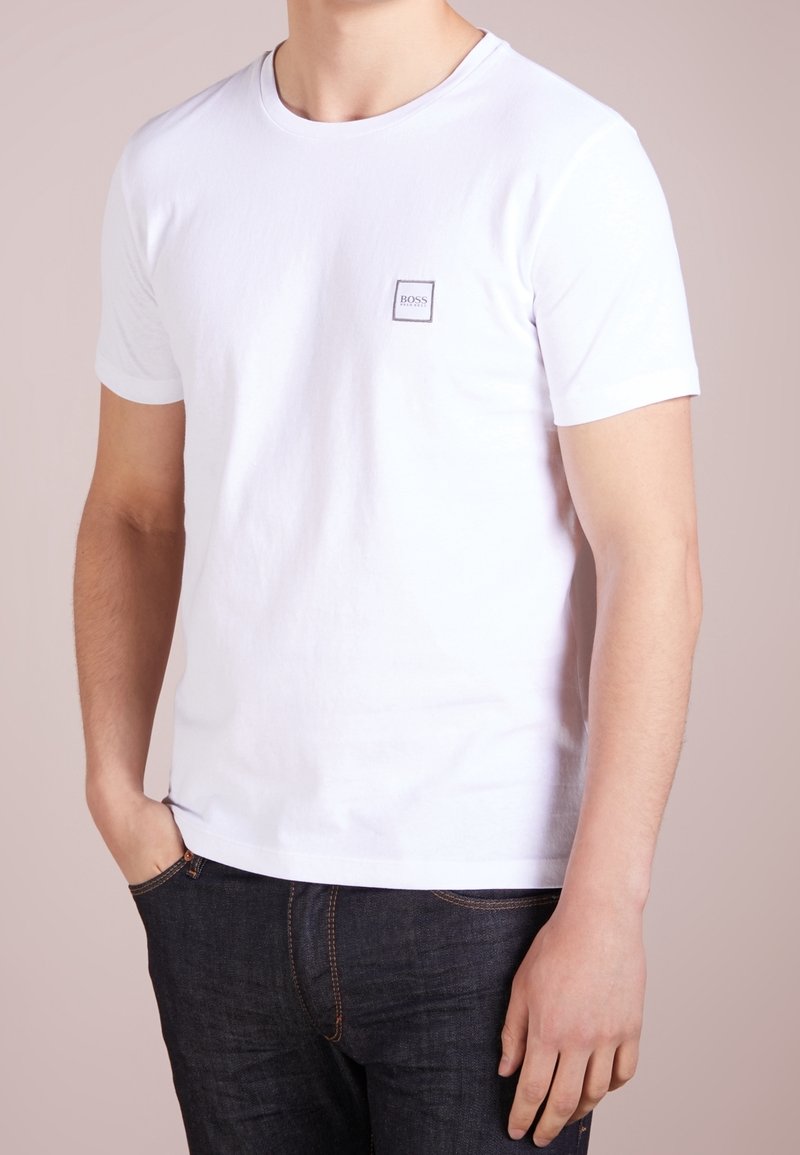 Homme portant un t-shirt blanc uni à manches courtes avec un petit logo "BOSS" sur la poitrine et un jean bleu foncé, les mains reposant de manière décontractée.