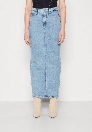 Vrouw draagt een lichtblauwe hoge taille denim midi-rok met een voornaad en beige enkellaarzen tegen een effen achtergrond.