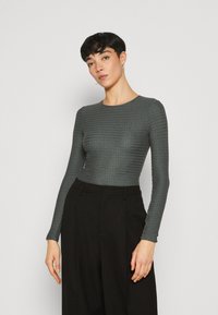 Langärmliges Oberteil aus strukturiertem, dunklem Graugewebe, mit Rundhalsausschnitt und taillierter Form, kombiniert mit hochgeschnittenen schwarzen Hosen.
