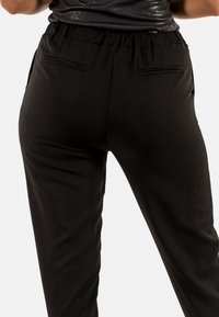 Persona que lleva pantalones de tiro alto negros con cinturilla elástica y dos bolsillos traseros tipo ojal, de pie contra un fondo claro y liso.