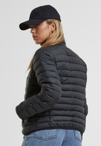 Veste noire rembourrée avec une texture matelassée, col haut et manches ajustées. Portée avec une casquette noire, accompagnée de jeans en denim bleu.