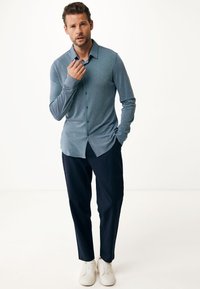 Ljust blå långärmad skjorta med krage, i kombination med marinblå byxor och vita sneakers, stående mot en neutral bakgrund.