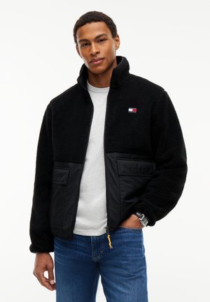 Junger Mann trägt eine schwarze Fleecejacke mit Fronttaschen, ein weißes T-Shirt, blaue Jeans und eine Armbanduhr, steht vor einem weißen Hintergrund.