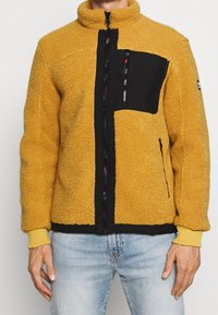 Giacca in pile di colore giallo senape con una tasca nera, chiusura con zip, colletto alto e polsini a coste. Superficie testurizzata e pannelli a contrasto.