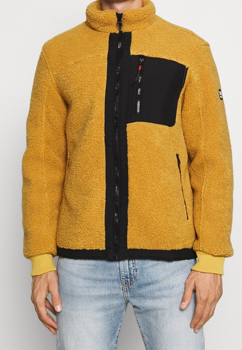 Giacca in pile di colore giallo senape con una tasca nera, chiusura con zip, colletto alto e polsini a coste. Superficie testurizzata e pannelli a contrasto.