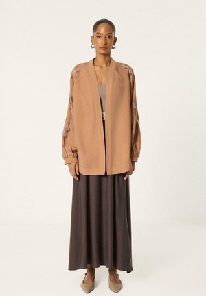 Femme portant une veste ouverte brodée marron clair sur un haut taupe et une longue jupe marron foncé, debout devant un fond uni.