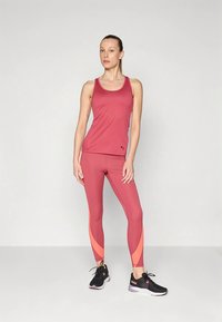 Γυναίκα που στέκεται με αυτοπεποίθηση φορώντας ροζ αθλητική μπλούζα και κολάν με κοραλλί λεπτομέρειες, και μαύρα αθλητικά παπούτσια Puma, σε απλό φόντο.