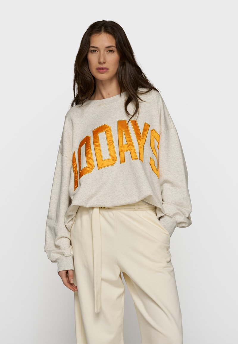 Femme aux longs cheveux bruns portant un sweat-shirt ample gris clair avec un grand texte orange « 100 DAYS », associé à un pantalon crème taille haute.