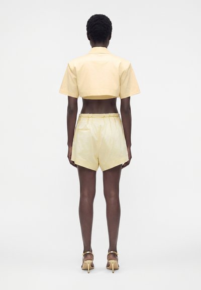 Femme portant une veste courte jaune pâle à manches courtes et un short taille haute assorti avec des sandales dorées à talons hauts, debout de dos.
