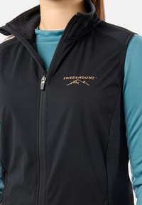 Czarna kamizelka softshell z wysokim kołnierzem, zamkiem z przodu oraz złotym logo z motywem góry. Nałożona na niebieską bluzkę z długim rękawem.