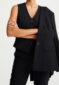 Czarna dopasowana kamizelka z dekoltem w serek, z trzema guzikami. Towarzyszy jej dopasowana, obszerna marynarka, opadająca na jedno ramię.